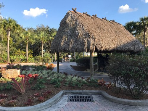 A thatched-roof gazebo surrounded by lush greenery and colorful plants in a sunny park setting.