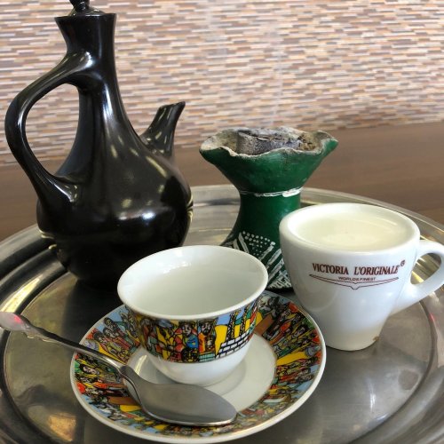A decorative tray with a black teapot, a green coffee pot, a cup of coffee, and a colorful saucer with a spoon.