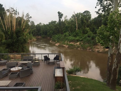 A serene river view from a deck with seating, surrounded by lush greenery and trees.