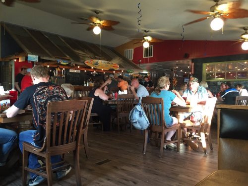 A lively restaurant interior with patrons dining at wooden tables, ceiling fans, and colorful decor.