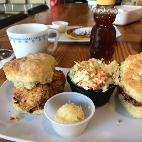 A plate with two chicken biscuits, coleslaw, and a small cup of butter, with syrup and a coffee cup in the background.