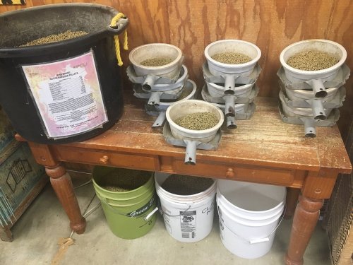 A wooden table with stacked containers of pet food and buckets underneath, against a wooden wall.