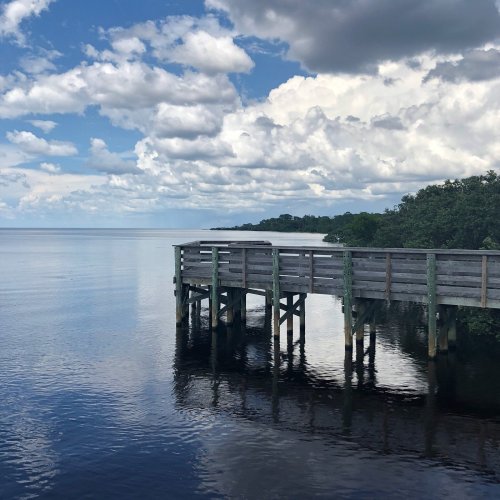 A wooden pier extends over calm water, reflecting clouds and greenery under a blue sky.