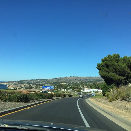 A scenic road curves through a coastal area with clear blue skies and hills in the background.