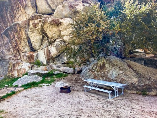 A picnic table and fire pit set against a backdrop of large rocks and sparse vegetation in a natural outdoor setting.
