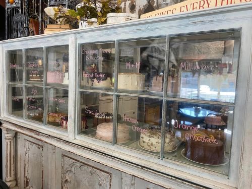 A display case filled with various cakes, each labeled with flavors like chocolate, peanut butter, and strawberry.