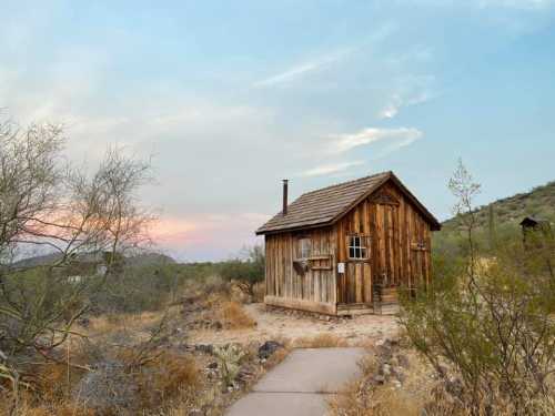 A rustic wooden cabin surrounded by desert vegetation under a colorful sky at dusk.