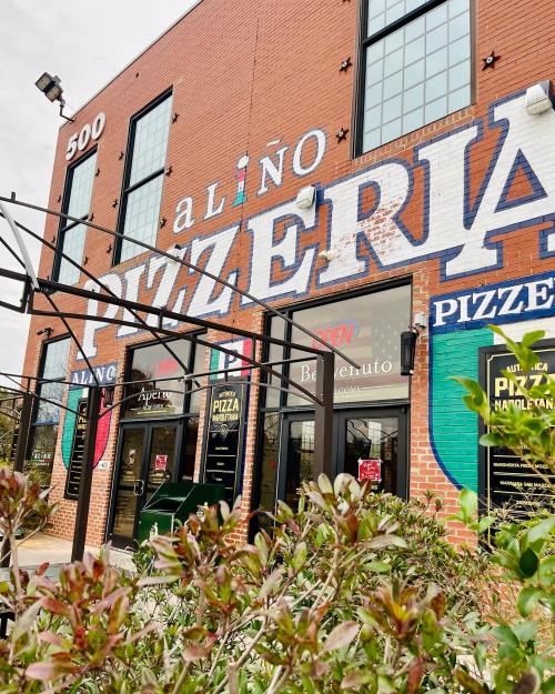 Exterior of a pizzeria with a large mural, featuring "Pizzeria" and "Open" signs, surrounded by greenery.
