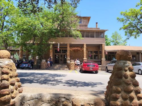 A stone building with a porch, surrounded by trees, with people and cars in front on a sunny day.