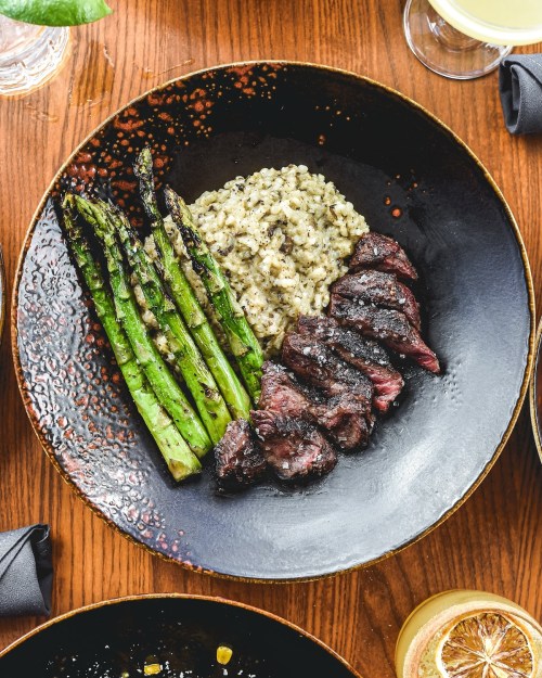 A plate featuring grilled asparagus, risotto, and sliced steak on a dark, textured plate.