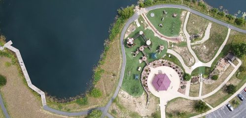 Aerial view of a park with playgrounds, walking paths, and a pond, surrounded by greenery and a parking area.