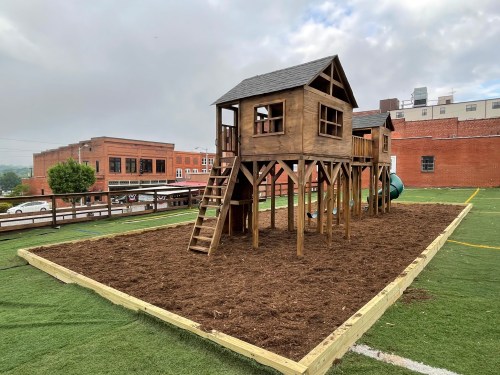 A wooden playground structure with two playhouses on stilts, surrounded by freshly laid mulch and grass.