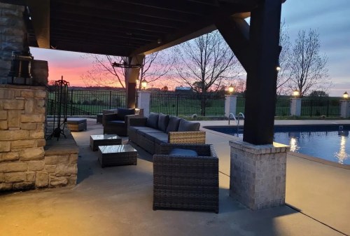 Cozy outdoor seating area by a pool at sunset, with a stone fireplace and trees in the background.