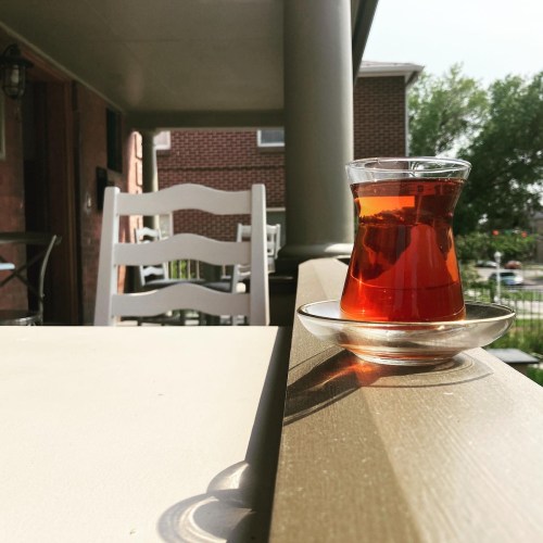 A glass of tea on a table, with a blurred background of chairs and greenery on a sunny day.