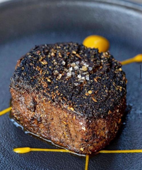 A perfectly cooked, seasoned steak on a black plate, garnished with yellow sauce and a sprinkle of salt.