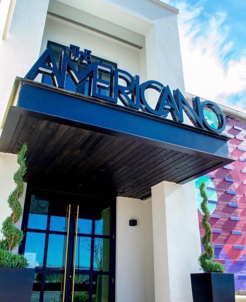 Signage for "The Americano" above a modern entrance, framed by decorative greenery and colorful wall patterns.