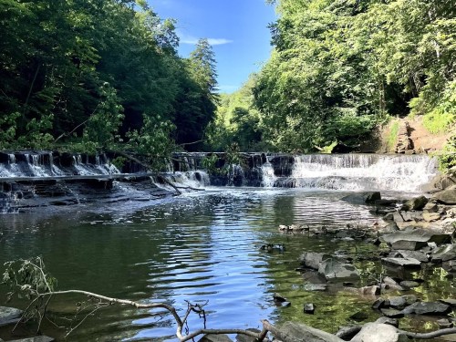 A serene waterfall cascades into a calm river, surrounded by lush green trees and rocky banks under a clear blue sky.