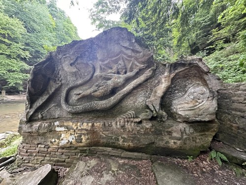 A carved rock featuring a dragon and other mythical creatures, surrounded by lush greenery near a river.