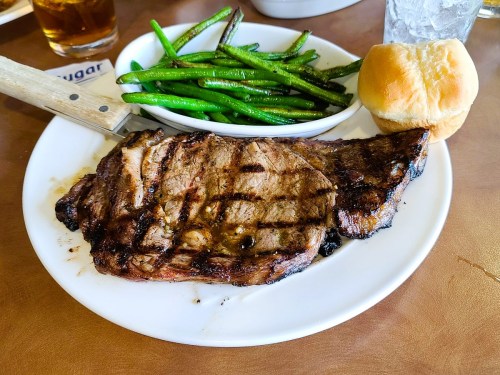 A grilled steak with grill marks, served with green beans and a dinner roll on a white plate.