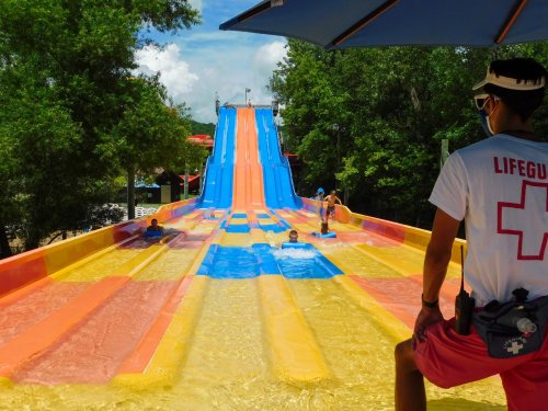 A lifeguard watches over a colorful water slide with people sliding down into a pool below, surrounded by trees.