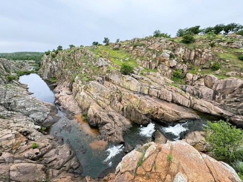 A rocky landscape with a winding river, surrounded by greenery under a cloudy sky.