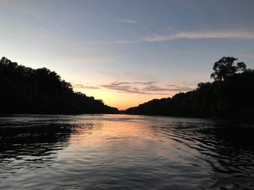 A serene river at sunset, reflecting colorful skies and surrounded by trees on both banks.