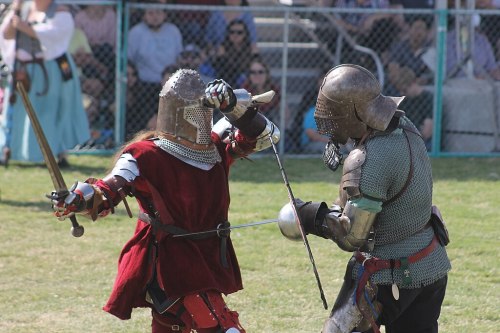 Two armored knights engage in a sword fight on a grassy field, with spectators watching in the background.