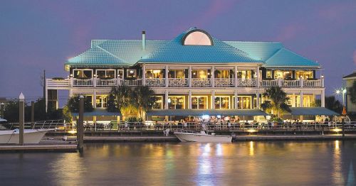A waterfront restaurant with a blue roof, illuminated at dusk, surrounded by palm trees and a calm waterway.