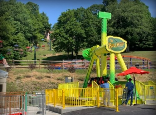 A colorful amusement ride called "The Bullwhip" with yellow barriers, set in a park with hills and other rides in the background.