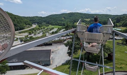 A person sits in a ferris wheel seat, overlooking a scenic landscape with hills and a winding road below.