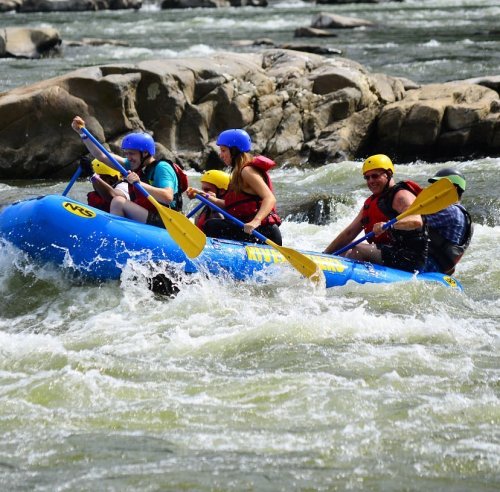 A group of six people in a blue raft navigating whitewater rapids, wearing helmets and life jackets.
