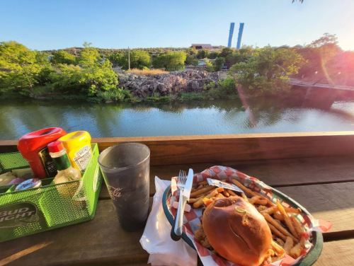A burger and fries on a table with condiments, overlooking a serene river and lush greenery.