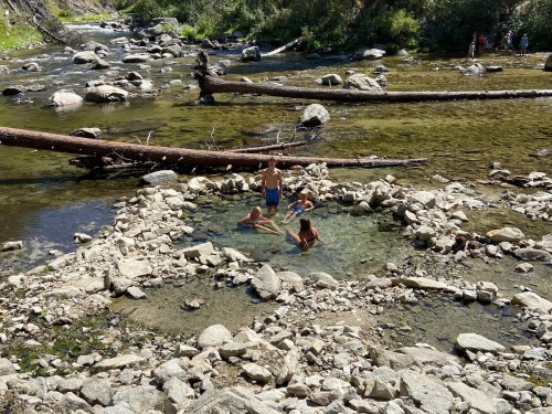 A natural hot spring surrounded by rocks, with people relaxing in the water and a river flowing nearby.