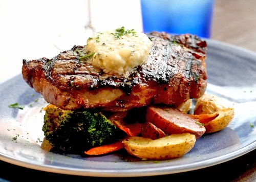 A grilled steak topped with herb butter, served on a plate with roasted vegetables and potatoes.