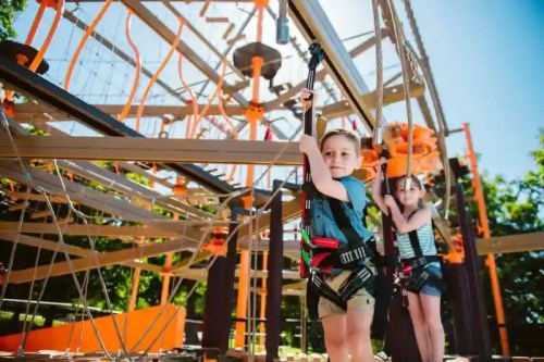 Two children in harnesses navigate a ropes course, surrounded by colorful structures and greenery on a sunny day.