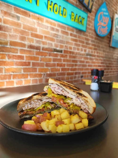 A close-up of a toasted sandwich filled with meats and veggies, served with seasoned potato cubes on a black plate.