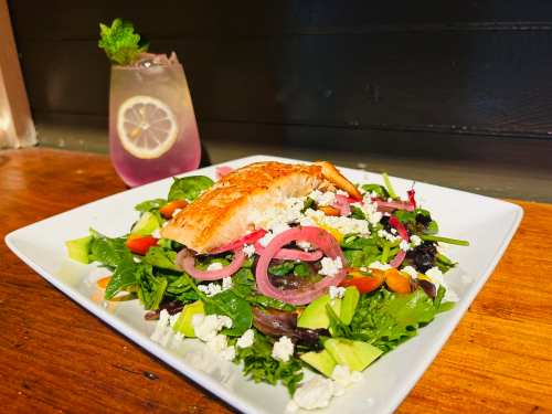 A vibrant salad topped with grilled salmon, mixed greens, cherry tomatoes, and feta, alongside a refreshing drink.