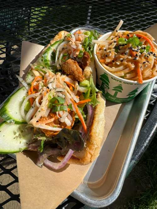 A sandwich with crispy chicken, fresh vegetables, and a side of noodles topped with sesame seeds on a metal tray.