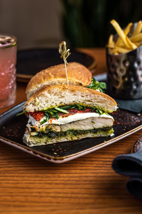 A sandwich with chicken, pesto, greens, and sun-dried tomatoes, served with a side of fries on a wooden table.