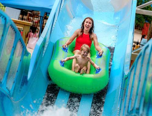 A woman and a boy joyfully slide down a water slide in a green inflatable tube, splashing into the water below.