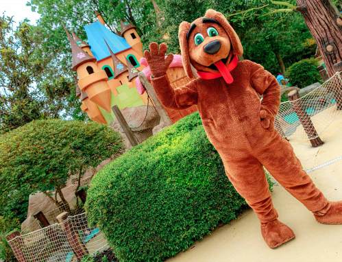 A cheerful cartoon dog mascot waves in front of a colorful castle in a park setting.