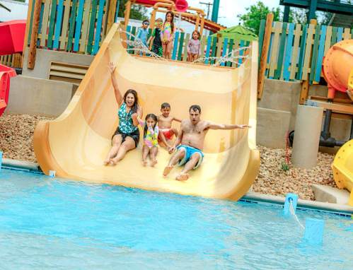 A family slides down a water slide into a pool, with children playing in the background at a water park.