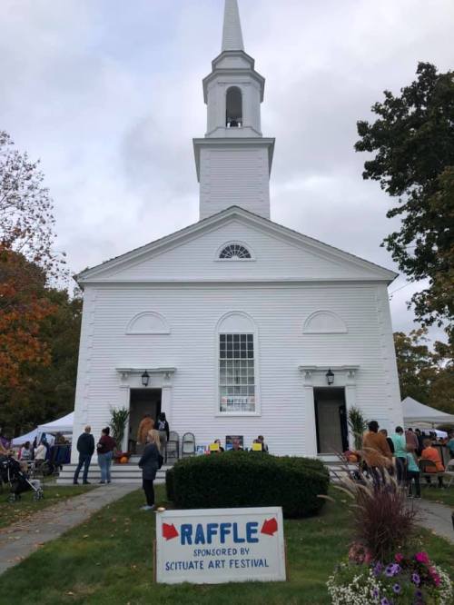 A white church with a tall steeple, surrounded by people and a sign for a raffle at the Scituate Art Festival.