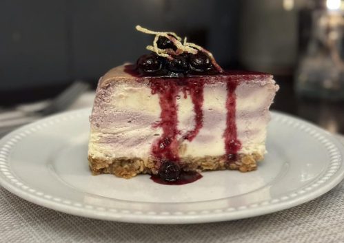 A slice of layered cheesecake topped with berry sauce and garnished with a twist of string, served on a white plate.