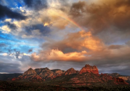 A vibrant sunset over red rock formations, with a rainbow arching through dramatic clouds in the sky.