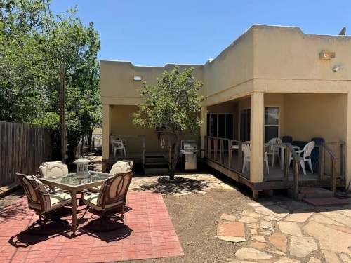 A sunny outdoor patio with a table and chairs, surrounded by trees and a house in a desert setting.
