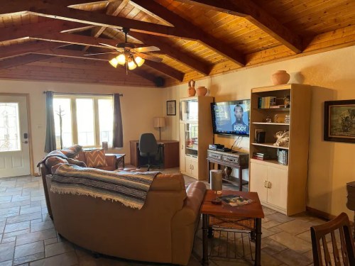 Cozy living room with wooden beams, a TV, bookshelves, and comfortable seating. Natural light from large windows.