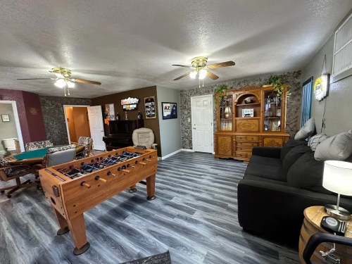 A cozy living room with a foosball table, seating area, and a piano, featuring light wood flooring and warm decor.