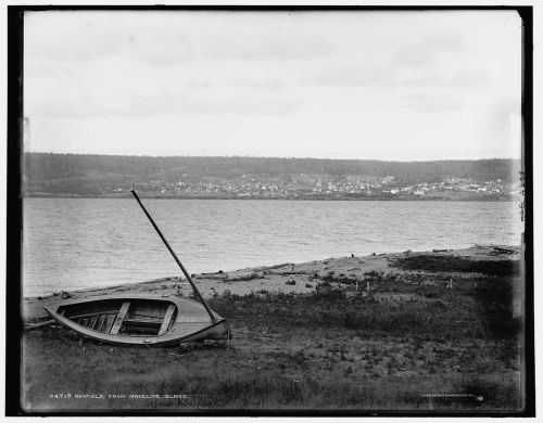 A weathered boat rests on the shore of a calm lake, with a distant town visible across the water under a cloudy sky.