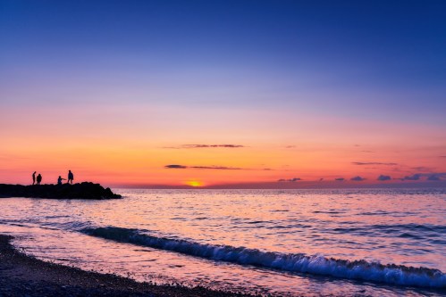 Silhouettes of people on a rocky shore at sunset, with vibrant colors reflecting on the calm ocean waves.